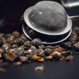 Boabe de cafea și frunze de ceai uscat așezate alături pe o masă de lemn, lângă o ceașcă și un ceainic