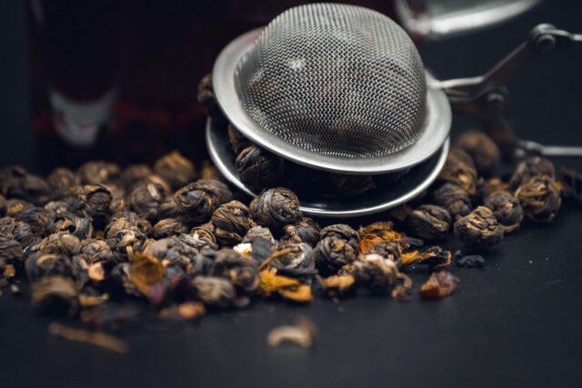 Boabe de cafea și frunze de ceai uscat așezate alături pe o masă de lemn, lângă o ceașcă și un ceainic
