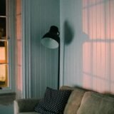Living luminat seara de o lampă cu abajur textil și lumină caldă, lângă o canapea cu perne