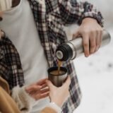 Termos metalic și o cană cu cafea aburindă pe o bancă din parc, într-o zi răcoroasă de toamnă