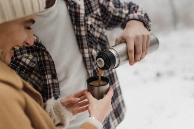 Termos metalic și o cană cu cafea aburindă pe o bancă din parc, într-o zi răcoroasă de toamnă