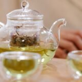 Ceainic din sticlă cu ceai verde limpede și o ceașcă mică alături, pe o tavă de lemn