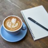 Caiet deschis cu un pix alături și o ceașcă de cafea pe o masă de lemn, lângă o fereastră luminoasă