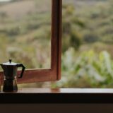 Ceașcă de cafea pe pervazul unei ferestre luminoase, lângă câteva plante verzi, într-o dimineață liniștită